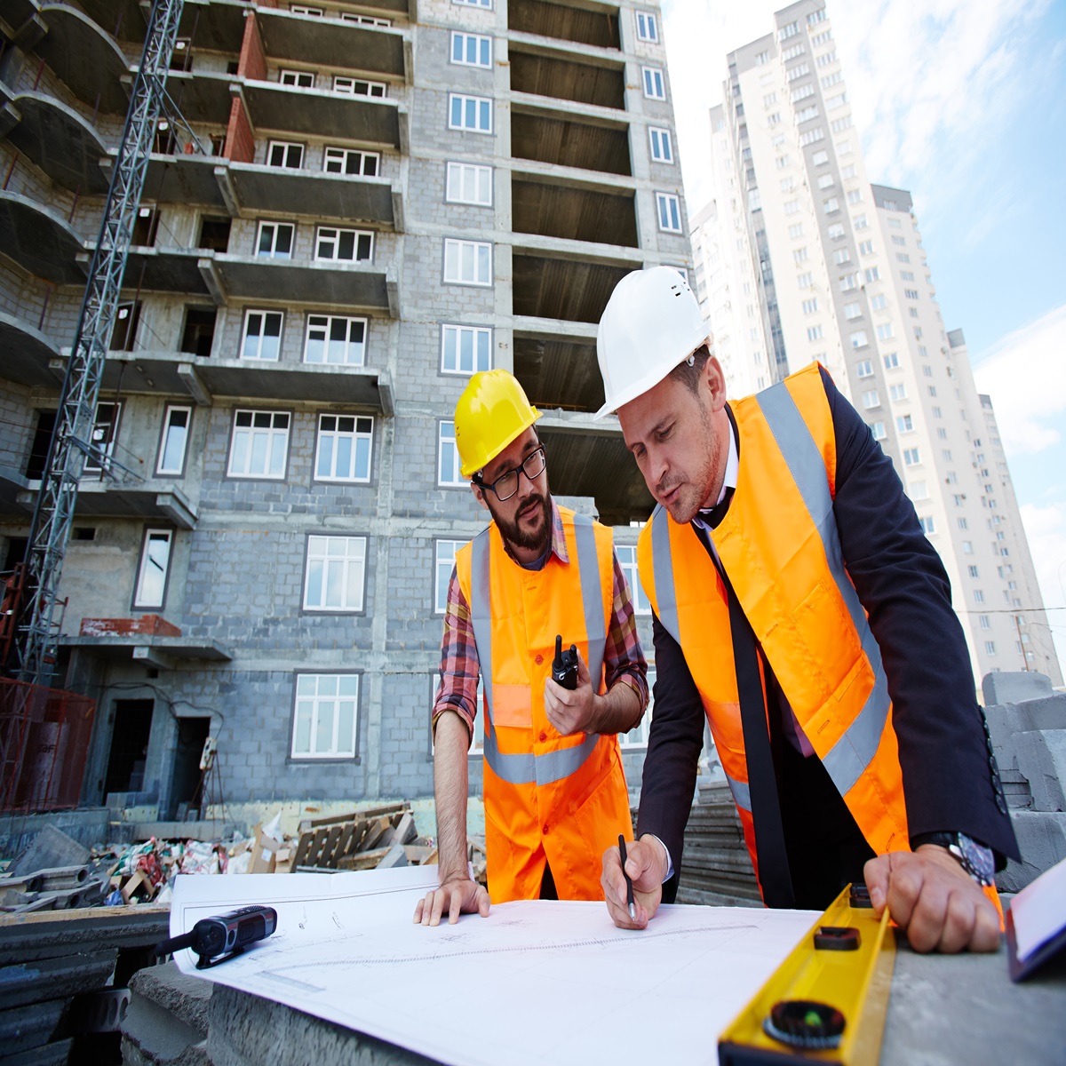 Two workers in hard hats and orange safety vests reviewing blueprints at a building construction site