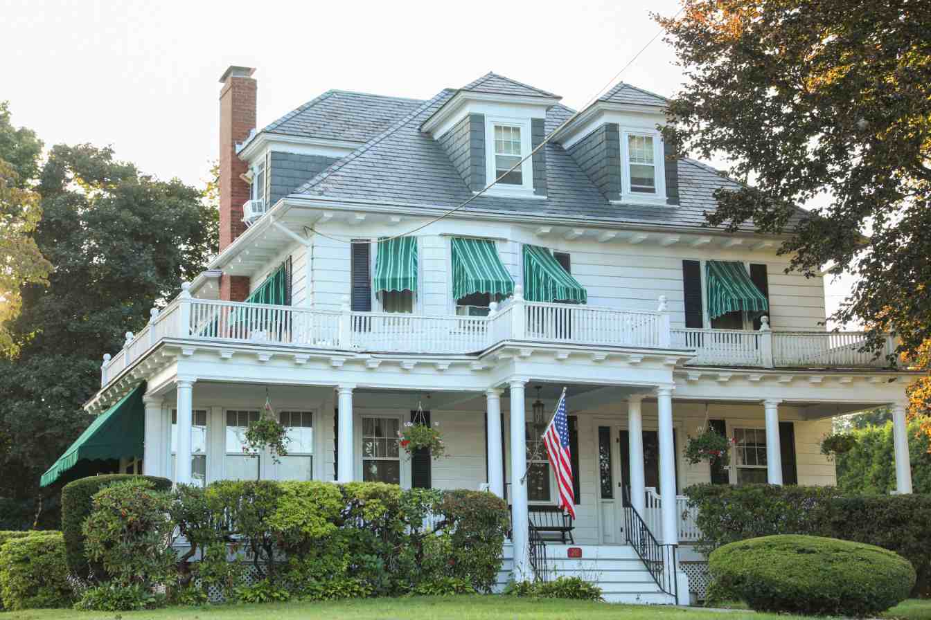 White two-story house with a wraparound porch, green striped awnings, dormer windows and US flag