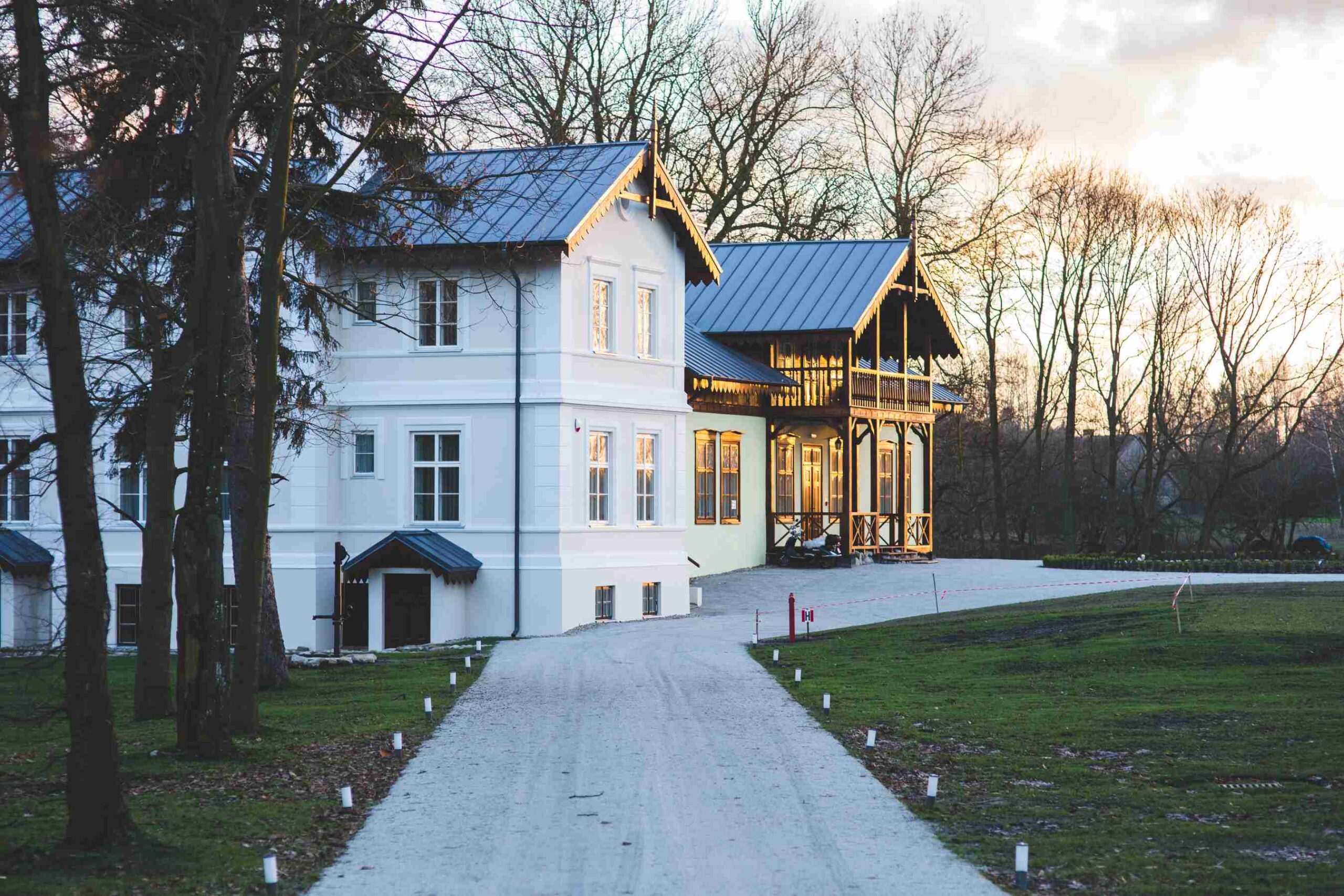 White two-story house with a dark metal roof and wooden gabled porch at the end of a gravel drive