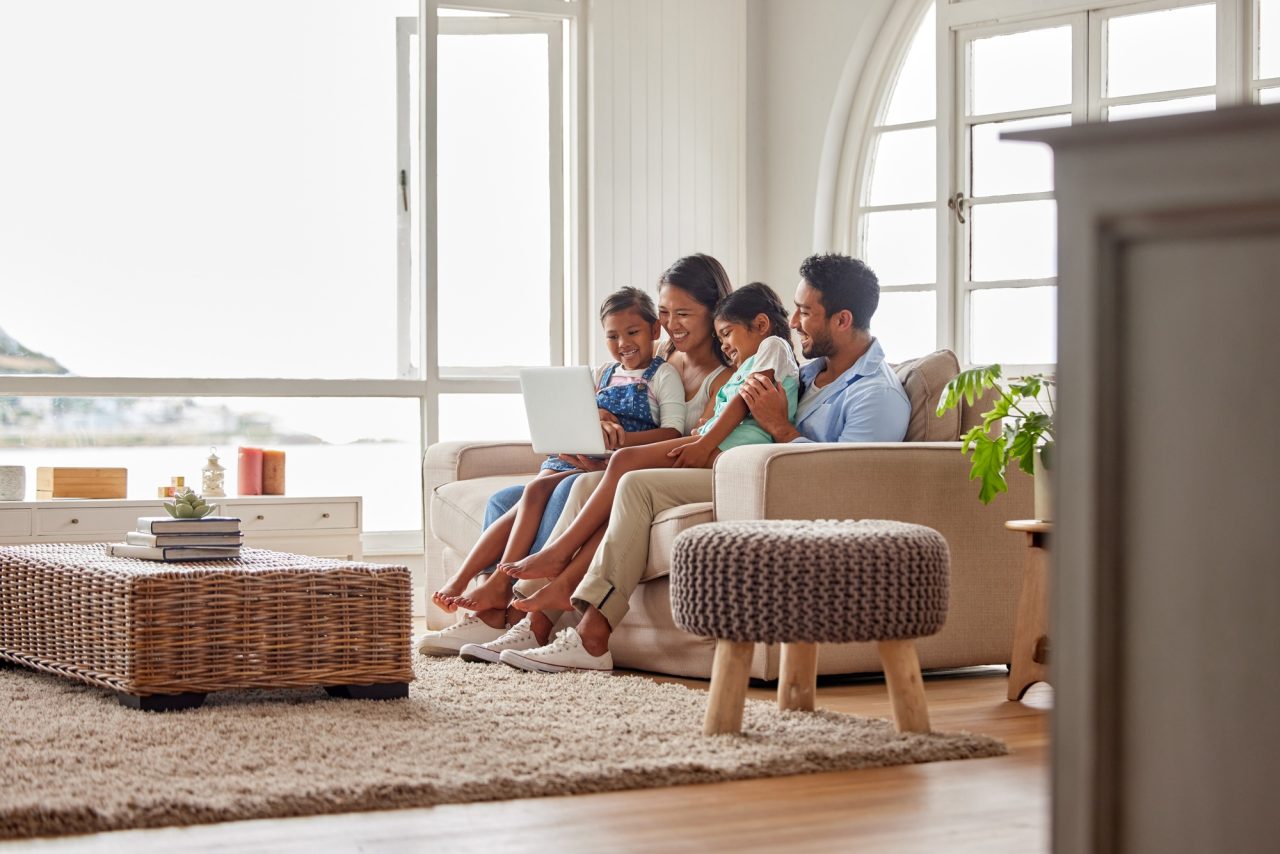 Family of four sitting on a beige sofa with a laptop in a bright living room with arched windows