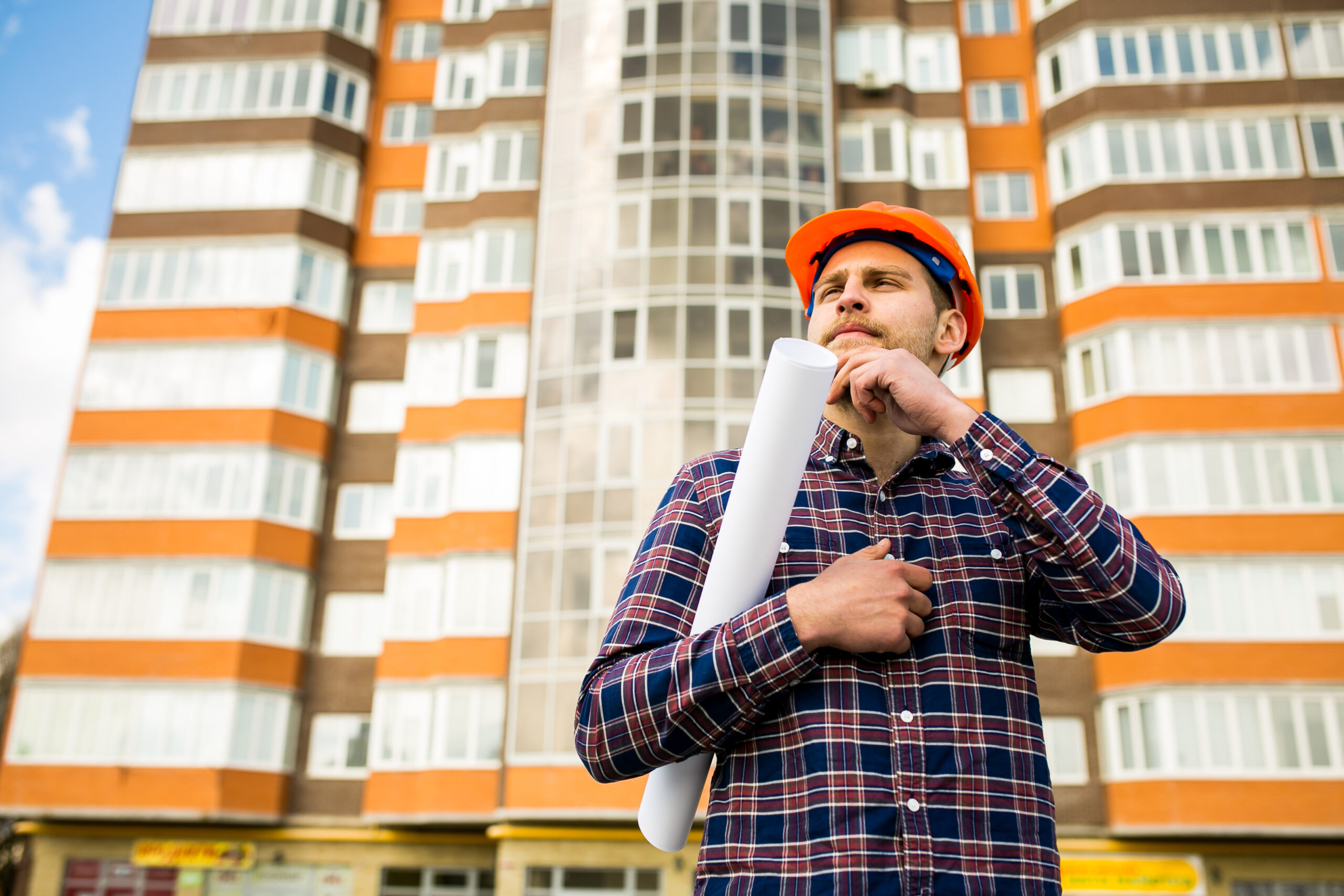 Worker in an orange hard hat holding rolled blueprints in front of a high-rise apartment building