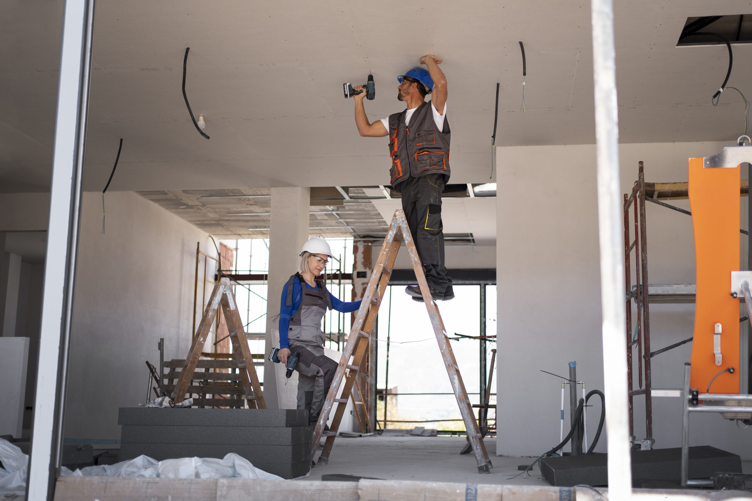 Two workers in hard hats fastening a drywall ceiling, one on a stepladder with a cordless drill