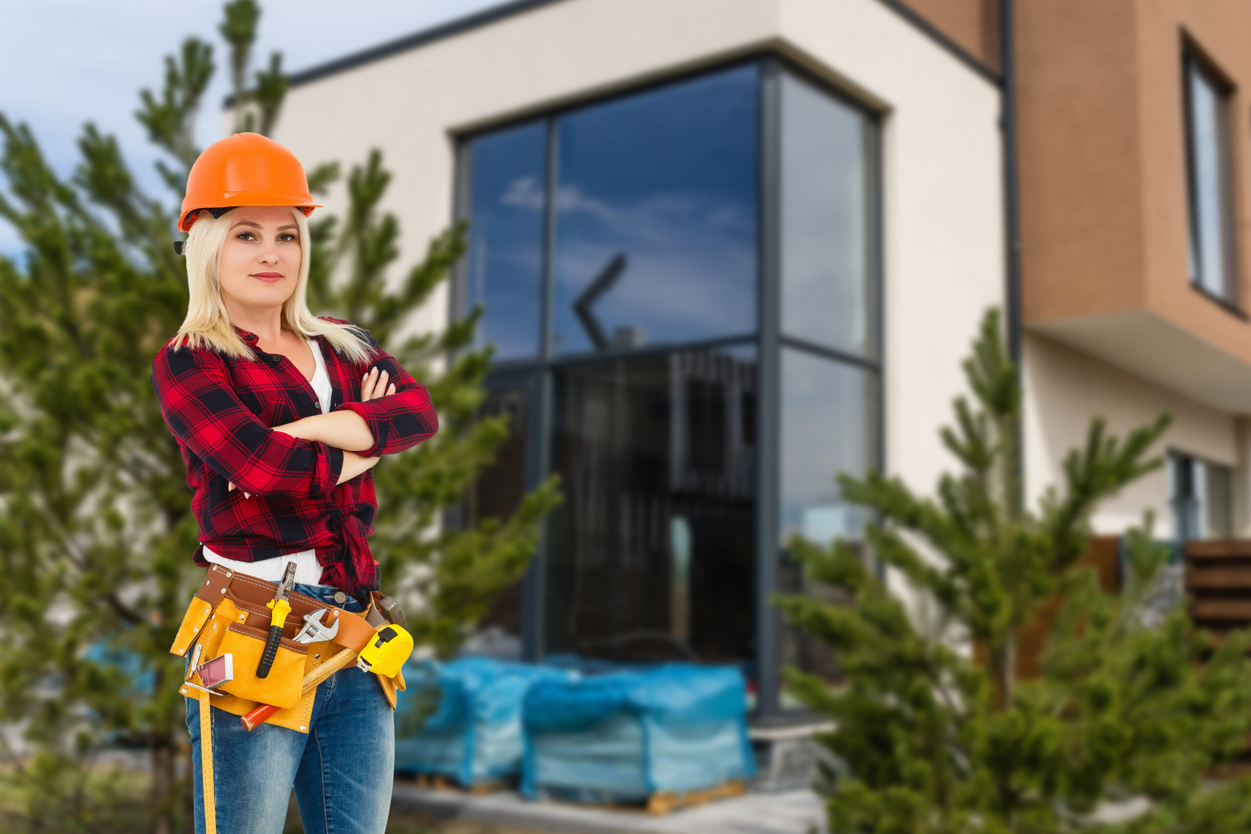Woman in an orange hard hat and leather tool belt standing with arms crossed outside a modern house