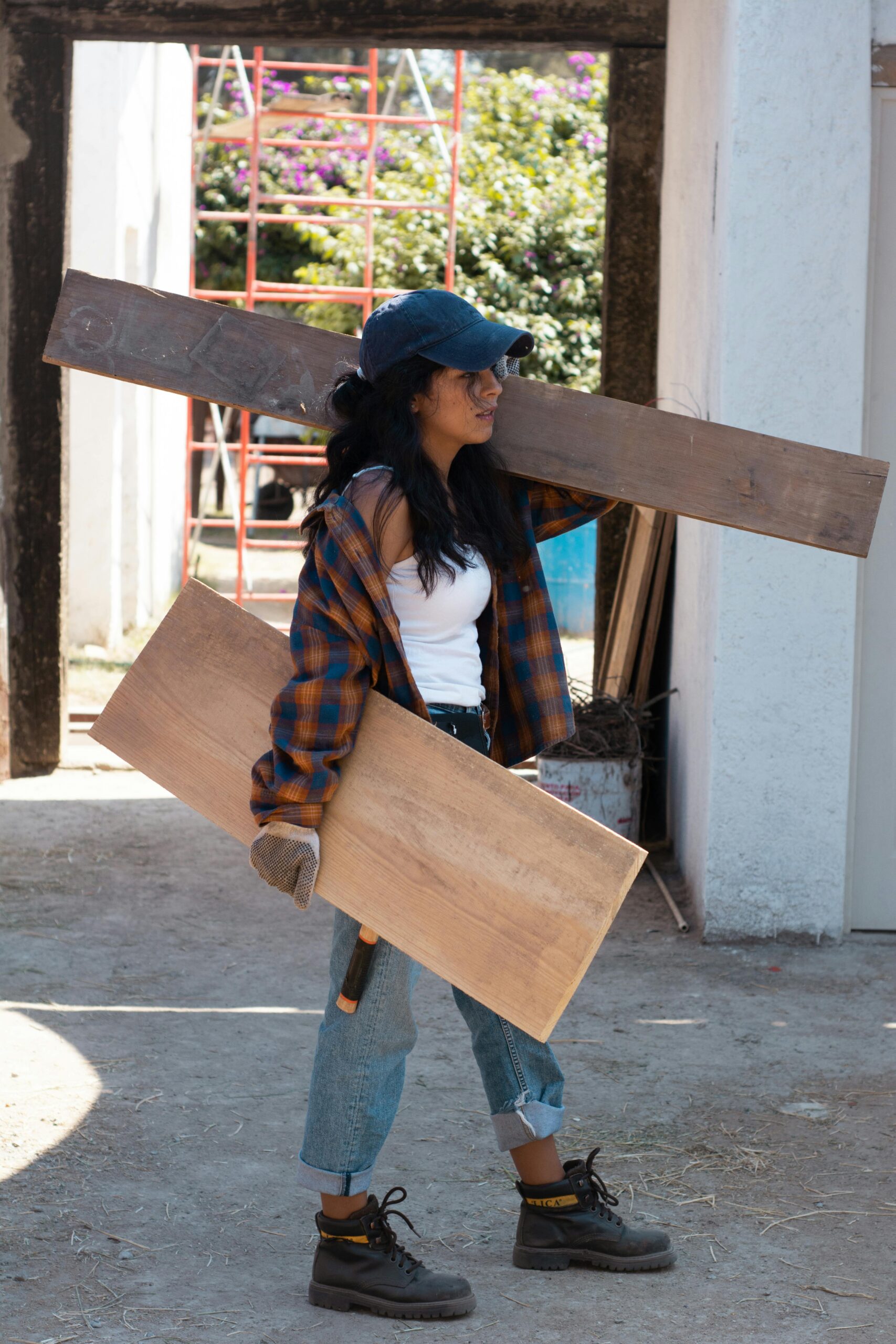 Worker in a flannel shirt and cap carrying two wooden boards at a construction site